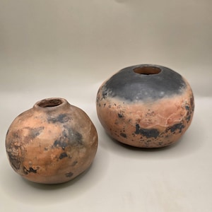 May include: Two spherical ceramic vases with small openings. The larger vase is black and peach, the smaller is peach and brown. The vases have a rustic, earthy appearance. The pottery appears handmade, with a natural, organic aesthetic.