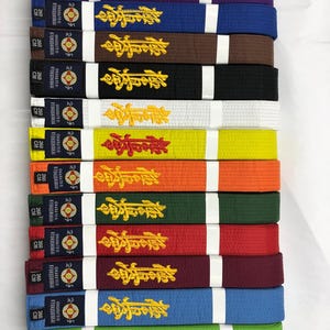 Puede incluir: Una pila de cinturones de karate en varios colores, incluyendo morado, azul, marrón, negro, blanco, amarillo, naranja, verde, rojo y burdeos. Cada cinturón tiene texto bordado en dorado y una sección blanca con texto y un emblema circular. Los cinturones miden 260 cm de largo.