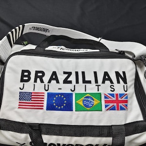 Puede incluir: Bolsa de deporte blanca y negra con las palabras "BRAZILIAN JIU-JITSU" en letras negras grandes. La bolsa presenta banderas bordadas de EE. UU., la UE, Brasil y el Reino Unido. Tiene una correa para el hombro y asas negras.