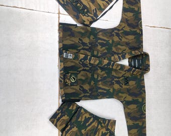 Puede incluir: Conjunto de uniforme de artes marciales con estampado de camuflaje, que incluye una chaqueta, pantalones y una bolsa con cordón. La tela presenta una mezcla de tonos verdes, marrones y negros. La chaqueta y los pantalones tienen parches con texto y logotipos. La bolsa con cordón tiene el texto "RVCA".