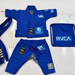 Op de afbeelding: Een blauwe vechtsport gi-set bestaande uit een jas, een broek met trekkoord, een blauwe riem met zwarte strepen en een bijpassende tas met trekkoord. De jas en broek zijn voorzien van witte RVCA- en VA-logo's en zwarte SHOYOROLL-labels.