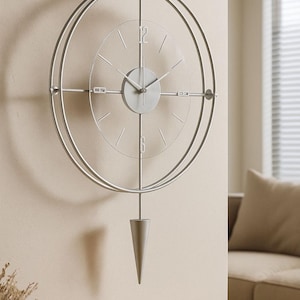 Pode incluir: Um relógio de parede prateado com um design minimalista. O relógio apresenta uma moldura circular, um mostrador transparente com marcadores de hora simples e um pêndulo. O relógio está montado numa parede bege, e a estética geral é moderna e elegante.