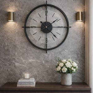 Puede incluir: Un gran reloj de pared redondo negro con un diseño minimalista cuelga de una pared de mármol gris. El reloj tiene manecillas negras y una esfera transparente. Dos apliques dorados flanquean el reloj. Debajo, un mueble de madera oscura contiene libros, una vela y un jarrón de rosas blancas.