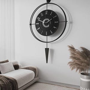 Op de afbeelding: Een grote, zwarte wandklok met een cirkelvormig ontwerp en Romeinse cijfers. De klok heeft een slinger en is aan een witte muur bevestigd. De diameter van de klok is ongeveer 61 cm.