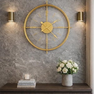 Puede incluir: Un reloj de pared circular dorado con una esfera transparente y números romanos. El reloj está montado en una pared gris con vetas de mármol, con dos apliques dorados a cada lado. Un ramo de rosas blancas se encuentra debajo.