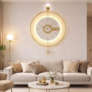 Könnte beinhalten: Eine moderne, goldgerahmte Wanduhr mit weißem Zifferblatt und goldenen Zeigern hängt über einem beigen Sofa. Die Uhr hat ein kreisförmiges Design mit einem leuchtenden Licht um das Zifferblatt. Der Raum ist in neutralen Tönen eingerichtet.