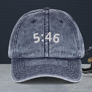 Puede incluir: Gorra de béisbol azul denim con el texto bordado "5:46" en blanco. La gorra tiene una visera curva y un aspecto vintage desgastado. Es un diseño clásico de seis paneles.