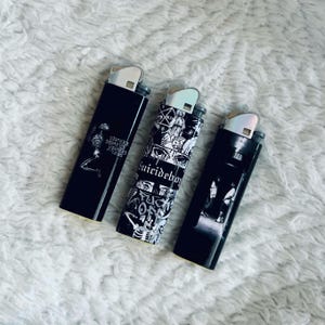 May include: Three black lighters with silver accents. One lighter features a skeleton graphic, another has a collage design with the text "Suicideboys", and the third has a grayscale image of people.