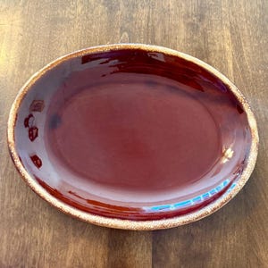 Pode incluir: Prato de servir oval em cerâmica com interior bordô profundo e brilhante. A borda tem uma borda texturizada marrom clara. O prato é provavelmente usado para servir alimentos.