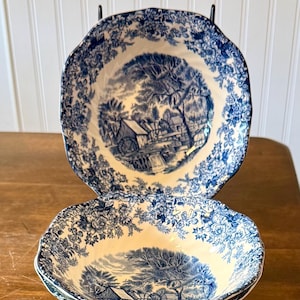 May include: Two blue and white ceramic bowls with a landscape design. The bowls have a scalloped edge and a detailed pattern of trees, buildings, and water features. The bowls are stacked on a wooden surface.