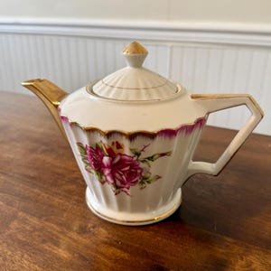 Op de afbeelding: Een witte porseleinen theepot met gouden details op de tuit, het handvat en het deksel. De theepot heeft een bloemenontwerp met roze rozen en groene bladeren. De rand is geaccentueerd met een dieproze band.