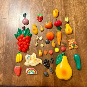 Może przedstawiać: Kolekcja kolorowych, plastikowych magnesów i elementów dekoracyjnych w kształcie owoców i warzyw. Zawiera kiść czerwonych winogron, żółtą gruszkę, truskawkę, marchewkę i misia. Przedmioty ułożone są na brązowej drewnianej powierzchni.