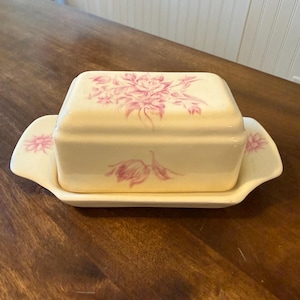 May include: Cream-colored ceramic butter dish with a rectangular lid and a matching base. The lid and base are adorned with pink floral patterns. The dish has a handle on each side of the base.