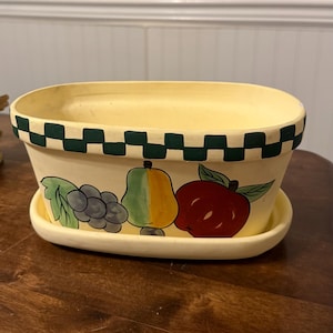 Könnte beinhalten: Ovale Keramik-Pflanzschale mit hellgelbem Boden und grün-weiß kariertem Rand. Die Pflanzschale zeigt handgemalte Illustrationen von Trauben, einer Birne und einem Apfel. Die Pflanzschale steht auf einer passenden Untertasse.