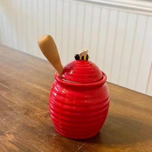 May include: A red ceramic honey pot with a wooden honey dipper. The pot has a lid with a small bee figurine on top. The pot is sitting on a wooden surface.