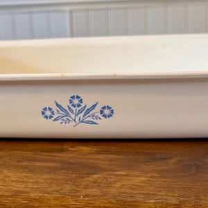 Puede incluir: Fuente rectangular blanquecina con esquinas redondeadas. La fuente presenta un diseño floral azul en el lateral. La fuente está sobre una superficie de madera.