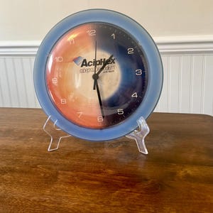 May include: A blue and orange clock with the text "AcipHex rabeprazole sodium" on the face. The clock is on a clear plastic stand and sits on a wooden surface.