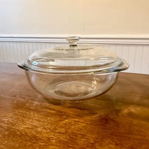 Puede incluir: Un cuenco de vidrio transparente con una tapa a juego. El cuenco tiene una forma redondeada con un borde ligeramente ensanchado y un asa a cada lado. La tapa tiene un asa de pomo y un borde decorativo.