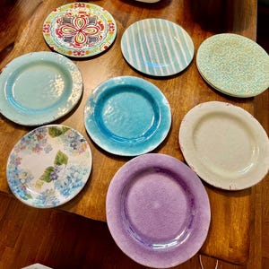 Puede incluir: Una colección de platos decorativos de cerámica en varios colores y diseños. Los platos incluyen diseños florales, a rayas y lisos en tonos de azul, verde, rojo y morado. Los platos están dispuestos sobre una superficie de madera.