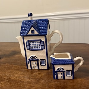 Può includere: Un set di teiera e lattiera in ceramica, progettato per assomigliare a case in miniatura. La teiera ha un tetto blu e pareti bianche con finiture blu, e una lattiera abbinata. Il set è posto su una superficie di legno.