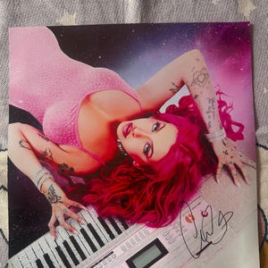 May include: A glossy photograph featuring a person with vibrant pink hair and a pink rhinestone-embellished top, lying across a white electronic keyboard. The background is a dark purple space nebula. A black ink signature is visible on the keyboard.