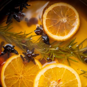 Può includere: Primo piano di una pentola piena di un liquido dorato, fette d'arancia, anice stellato, rametti di rosmarino e un bastoncino di cannella. Gli ingredienti sono immersi nel liquido, creando un'immagine calda. La scena suggerisce una bevanda speziata o una preparazione culinaria.
