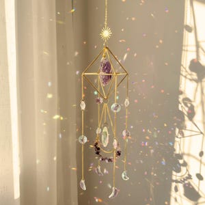Op de afbeelding: Een hangende zonnevanger met een geometrisch gouden frame met een paarse kristal. Heldere en paarse kralen en maansikkels hangen aan het frame. Een gouden zonnetje bekroont het ontwerp, wat een decoratief effect creëert.