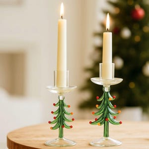 Puede incluir: Dos candelabros de vidrio con forma de árbol de Navidad con velas crema encendidas. Las bases verdes de los árboles tienen detalles rojos y naranjas. Los candelabros están sobre una superficie de madera, con un árbol de Navidad al fondo.
