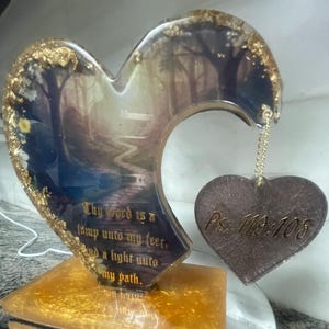 May include: A heart-shaped resin art piece with a forest scene, gold accents, and the text "Thy Word is a lamp unto my feet, and a light unto my path." A smaller heart pendant is attached, inscribed with "Ps. 119:105".