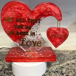May include: A red heart-shaped resin sculpture with the words "Let all that you do be done in Love" and "1 Cor 16:14". The sculpture has a small heart charm hanging from it and sits on a red base with confetti.