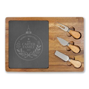 May include: A wooden cheese board with a dark gray slate insert. The slate is engraved with a Christmas ornament design and the text "MERRY CHRISTMAS THE CARTER FAMILY 2025". Three cheese knives with wooden handles are included.