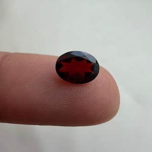 Può includere: Una gemma ovale, sfaccettata, di colore rosso scuro. La gemma presenta un motivo a forma di stella visibile al suo interno. La pietra è tenuta su un dito contro uno sfondo chiaro e semplice. Le sfaccettature catturano la luce.