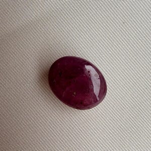 Könnte beinhalten: Ein polierter, oval geformter Edelstein in tiefem Burgunderrot. Der Stein hat eine glatte, glänzende Oberfläche mit subtilen inneren Einschlüssen. Der Edelstein ist vor einem neutralen, strukturierten Hintergrund platziert, der seine satte Farbe und Form hervorhebt.
