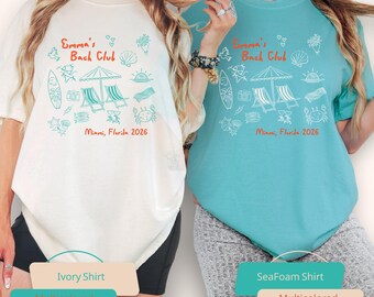 May include: Two t-shirts, one ivory and one seafoam, each with a beach-themed design in multiple colours. The design includes the text "Emma's Bach Club" and "Miami, Florida 2026", plus illustrations of beach chairs, a surfboard, and other beach elements.