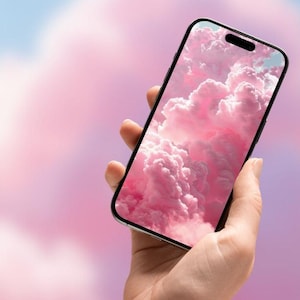 May include: A black smartphone displays a pink cloud wallpaper. The phone is held in a hand against a soft, blurred background of pink and blue hues. The screen shows fluffy, pink clouds.