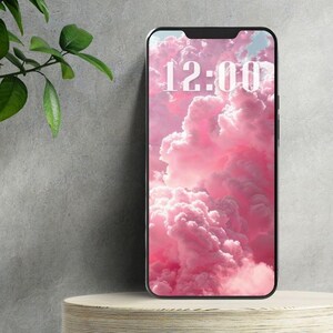 May include: A smartphone displays a pink cloud wallpaper with the time 12:00 in white text. The phone rests on a light-colored, round wooden stand. Green leaves are visible on the left side of the image.