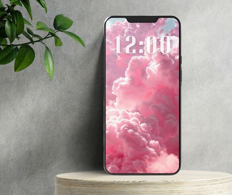 May include: A smartphone displays a pink cloud wallpaper with the time 12:00 in white text. The phone rests on a light-colored, round wooden stand. Green leaves are visible on the left side of the image.