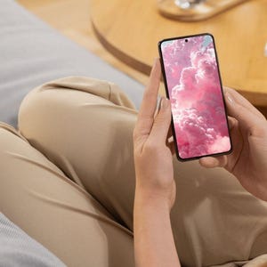 May include: A black smartphone with a pink cloud wallpaper is held by a person. The phone's screen displays a vibrant image of fluffy, pink clouds. The person is wearing tan pants and sitting on a gray surface.