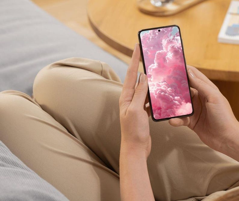 May include: A black smartphone with a pink cloud wallpaper is held by a person. The phone's screen displays a vibrant image of fluffy, pink clouds. The person is wearing tan pants and sitting on a gray surface.
