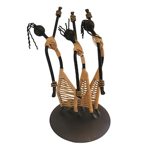May include: A decorative metal sculpture featuring three stylized figures with black bodies and heads. The figures are adorned with woven tan material and stand on a dark brown circular base. The sculpture has a unique, handcrafted appearance.