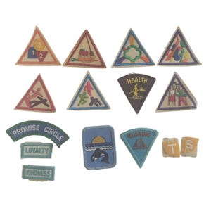 May include: A collection of vintage Girl Scout patches in various shapes and designs. Patches include triangular, rectangular, and arched shapes with embroidered images and text. Colors include red, green, blue, and yellow.