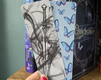 May include: A hand holds three bookmarks. One features a sword design with smoke, another has a blue butterfly pattern, and the third has the word "Nimerelle". The bookmarks are on a wooden shelf with books.