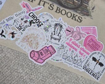 Puede incluir: Un conjunto de pegatinas blancas con varios diseños y textos, incluyendo frases como "Violent little thing" e "I didn't choose violence." Las pegatinas presentan ilustraciones de murciélagos, corazones y un trono.