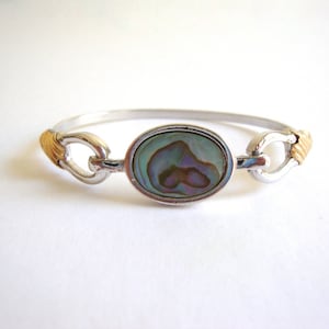 May include: Silver-tone bangle bracelet with gold-tone accents and an oval abalone shell centerpiece. The bracelet features a hinged design with a secure clasp. The abalone shell displays iridescent blues, greens, and purples.
