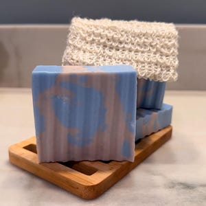 Puede incluir: Una pastilla de jabón rectangular con un patrón jaspeado en azul claro y rosa pálido, sobre una jabonera de bambú con listones. Un paño exfoliante tejido de color blanco roto está doblado y colocado directamente sobre el jabón.