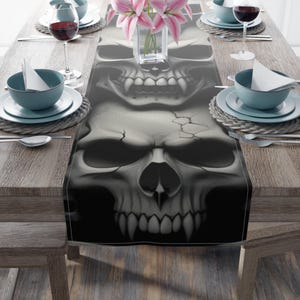 May include: A rectangular table runner featuring a repeating grayscale skull pattern with visible cracks on the cranium and sharp teeth. The runner is centered on a wooden table set with blue bowls, white napkins, silverware, and wine glasses.