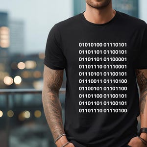 May include: Black t-shirt with white binary code printed vertically. The code appears to be a series of ones and zeros. The shirt is a crew neck and is being worn by a person. The background is blurred with city lights.
