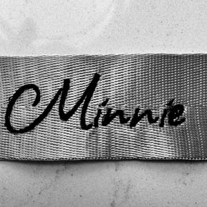 May include: Close-up of a silver woven ribbon with the name "Minnie" embroidered in black cursive script. The ribbon has a textured appearance and is set against a light gray background.