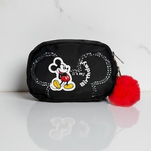 May include: Black zippered pouch featuring a Mickey Mouse graphic and the text "It's my Birthday!". The pouch has a red pom-pom attached to the zipper and is decorated with sparkling accents. The pouch is designed with a rounded shape.
