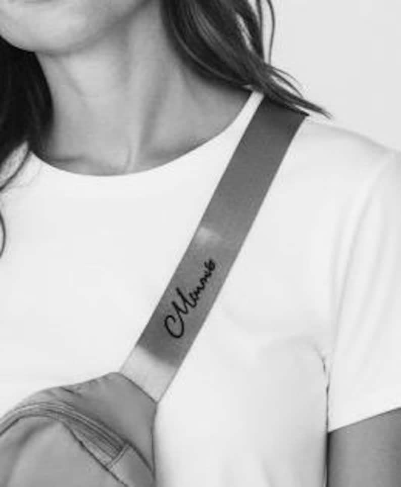 May include: A close-up of a person wearing a white t-shirt with a gray fanny pack. The fanny pack strap has the word "Mommy" in cursive script. The strap is across the person's shoulder.